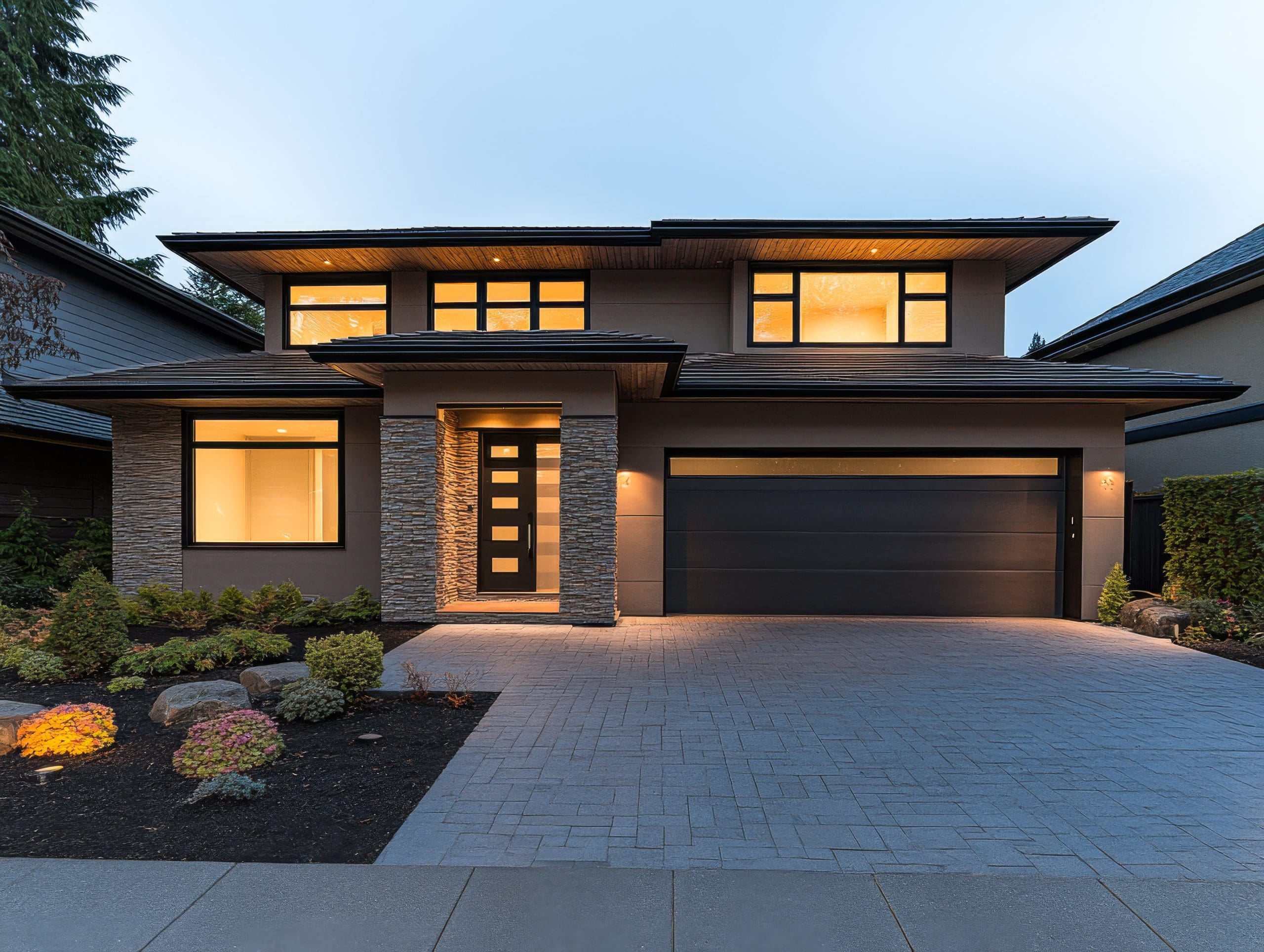 A modern two-story house with stone accents, large windows, and a black garage door. The driveway is paved with light gray bricks. Soft lighting from inside the house highlights the well-maintained front garden with shrubs and rocks.