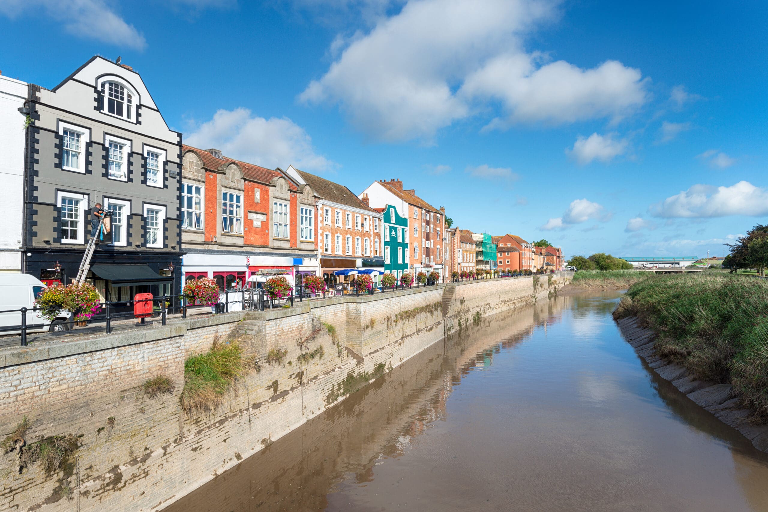 A calm river flows through a cityscape with colorful buildings lining the waterfront under a blue sky. The structures are mostly brick, with vibrant facades and multiple windows, creating a picturesque urban scene.