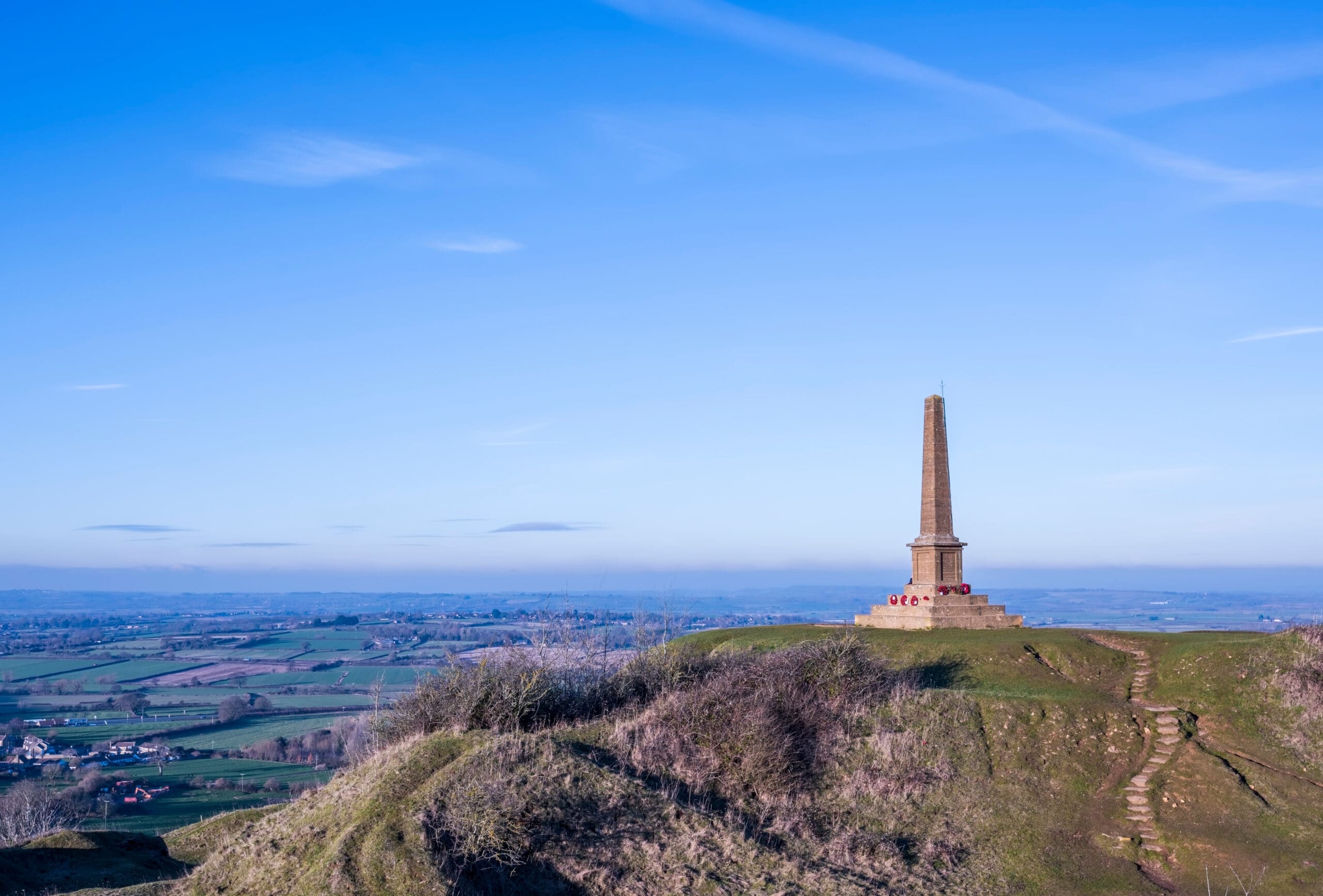 A tall obelisk monument stands atop a grassy hill under a clear blue sky. The landscape stretches out below with lush green fields and a winding path leading to the monument.