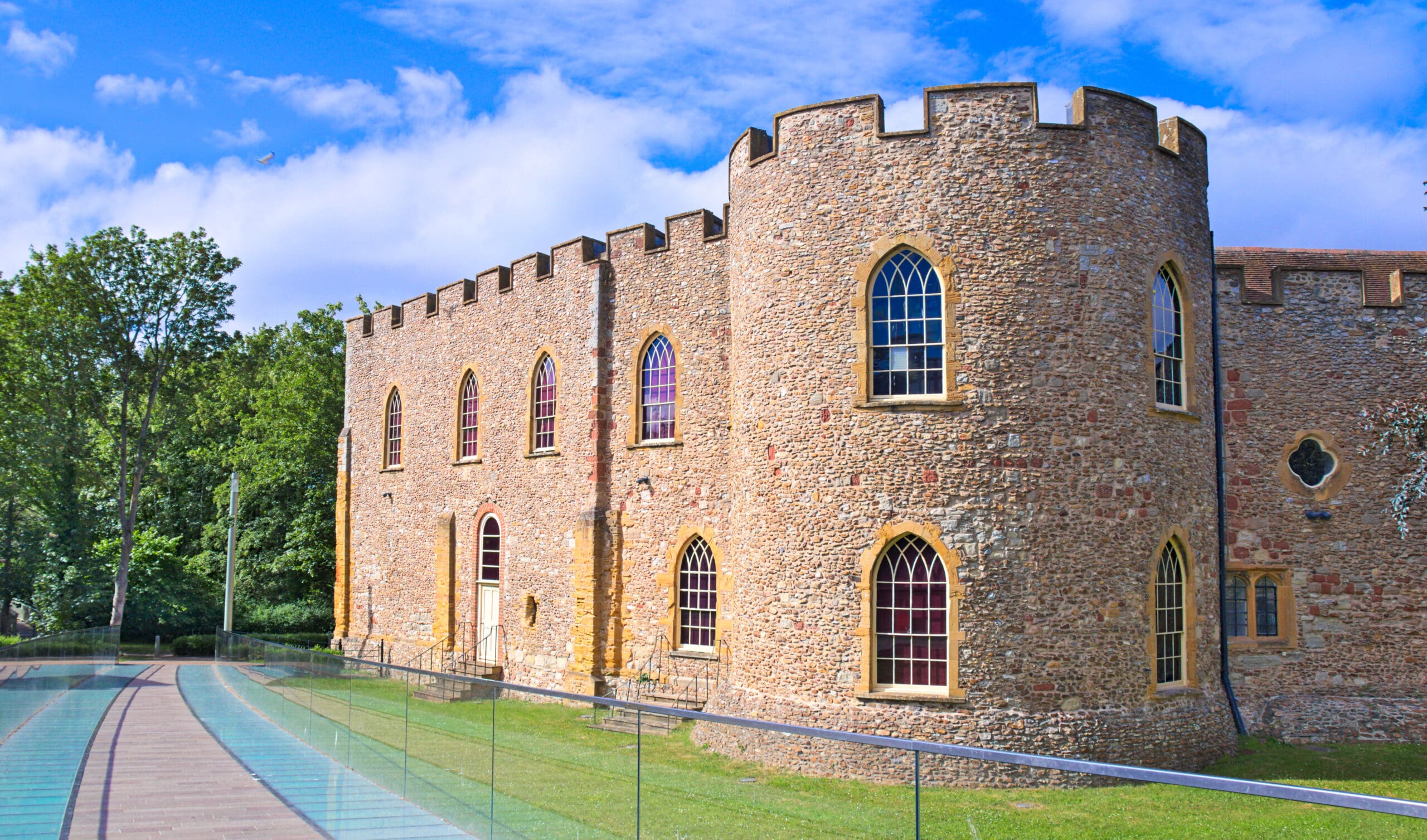 A stone castle with arched windows and a crenelated roof stands beside a glass walkway. The building is surrounded by lush greenery under a blue sky with scattered clouds.