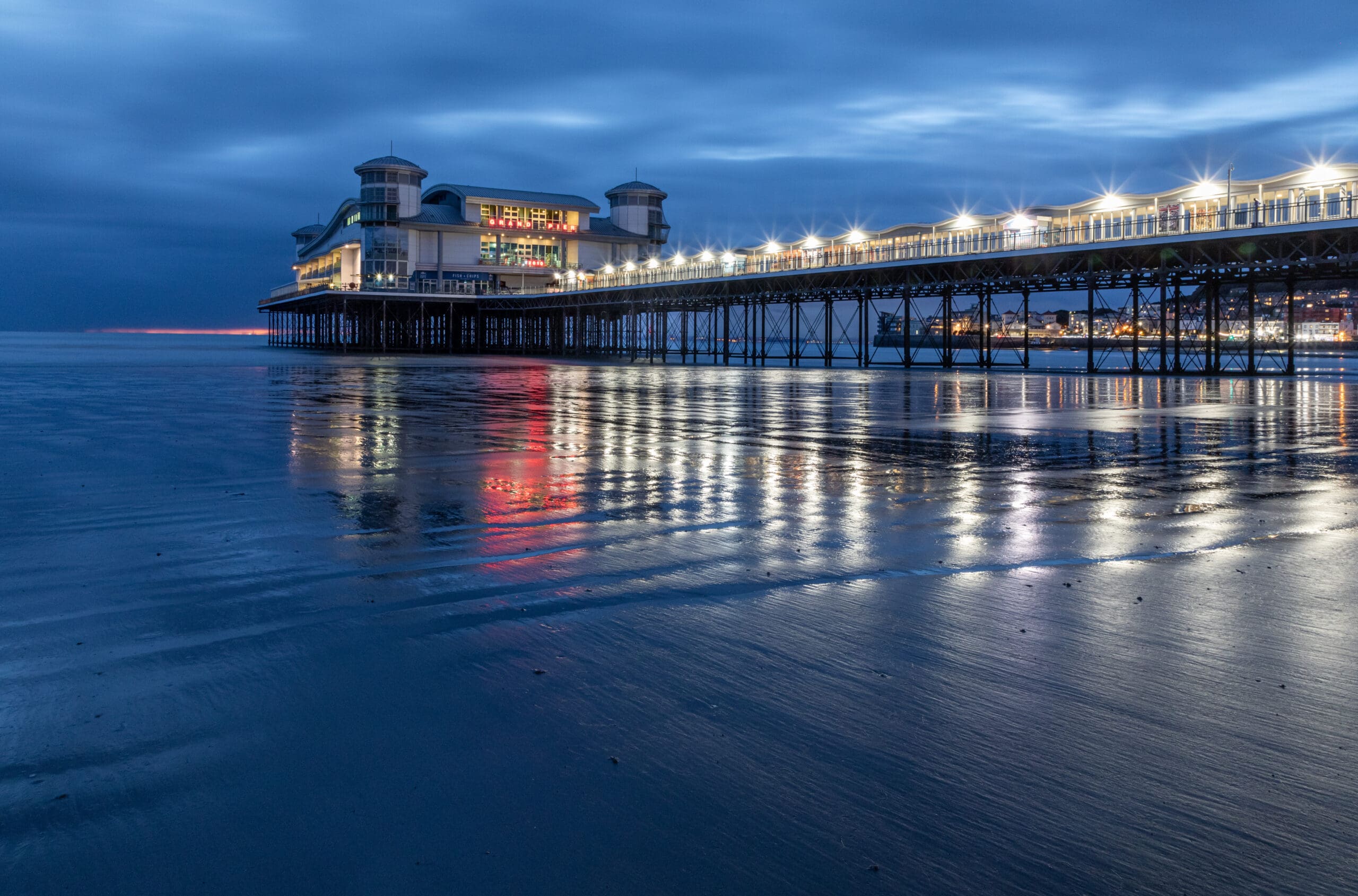 A pier with a building extends over the calm sea at dusk, illuminated by rows of bright lights. The water reflects the lights, creating a shimmering effect. The sky is a deep blue with scattered clouds, and the sandy beach is partially visible.