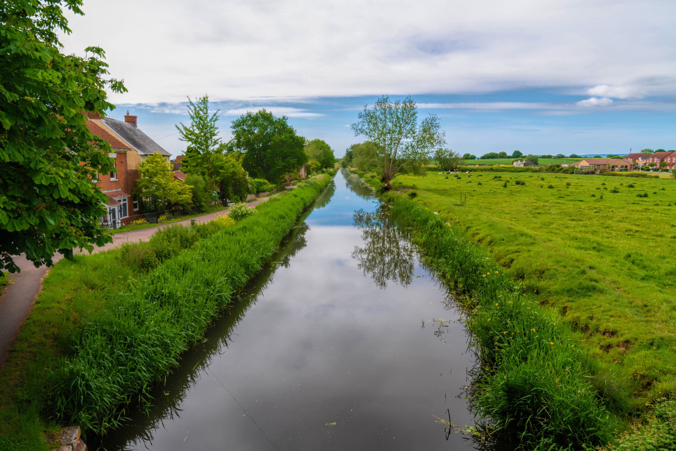 A serene rural scene with a narrow, calm canal flanked by lush green grass and trees. A red-brick house is visible on the left, and fields stretch into the distance under a partly cloudy blue sky.