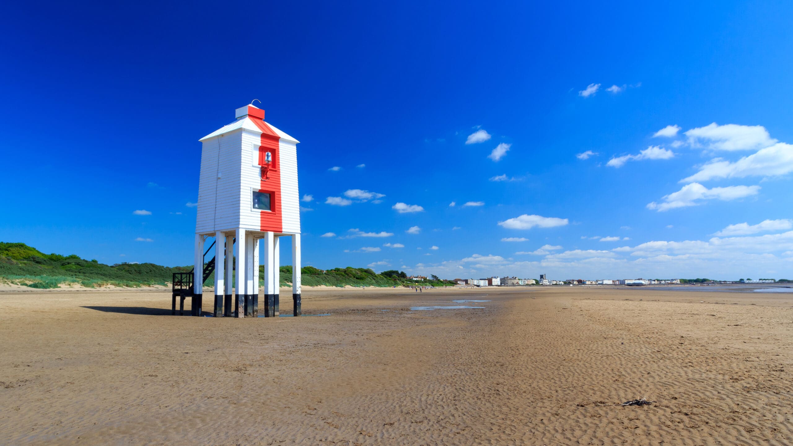 A white and red lighthouse stands on stilts on a sandy beach under a bright blue sky. The shoreline is visible in the background with green vegetation and distant buildings. Scattered clouds dot the sky.