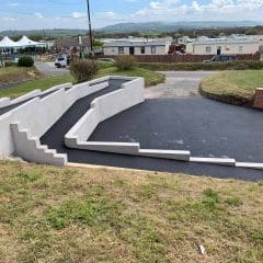 A newly paved driveway with white, modern, angular retaining walls on a sloped yard, near a residential area with houses, cars, and a view of distant hills under a partly cloudy sky.