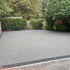 A freshly laid, smooth grey resin driveway bordered with dark bricks, surrounded by stone walls, green plants, and trees in a residential garden.