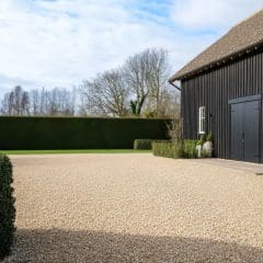 A modern black barn with large double doors stands beside a gravel courtyard, bordered by neatly trimmed hedges and surrounded by leafless trees under a partly cloudy sky.