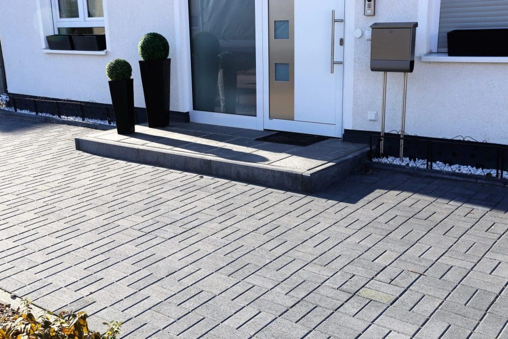 Modern house entrance with a light gray door, two tall black planters with round topiary shrubs, a gray mailbox, and a brick-paved driveway in a geometric pattern.