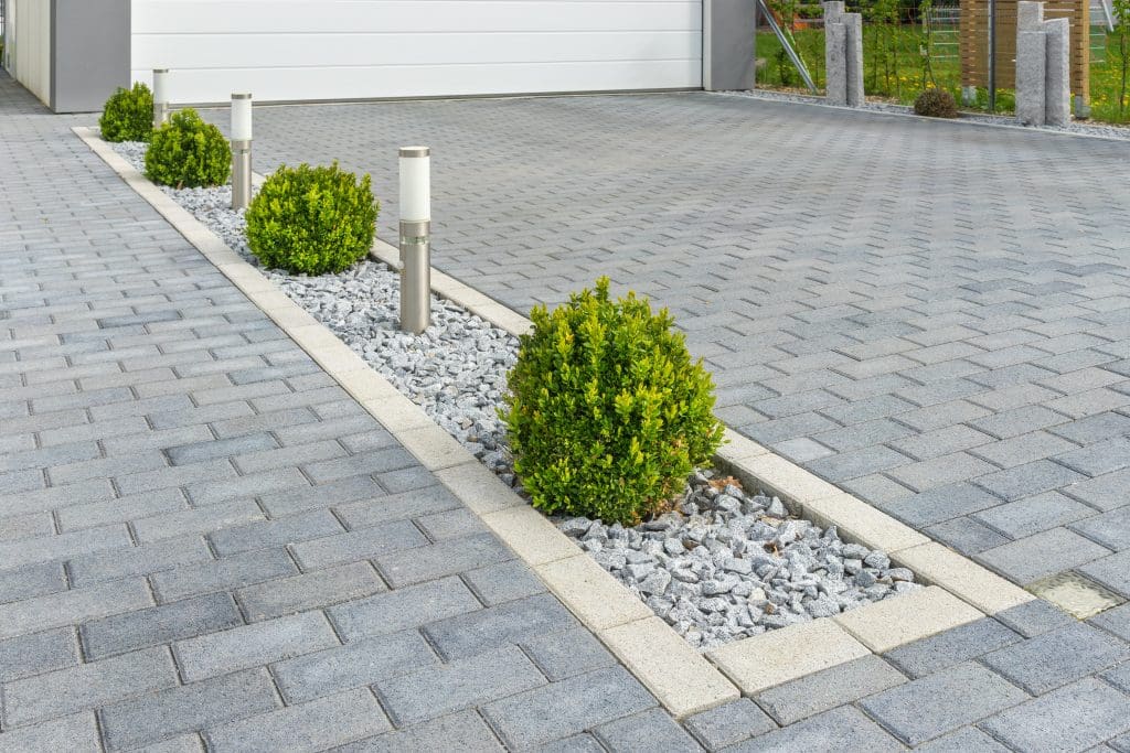 A modern driveway with gray paving stones, featuring a narrow landscaped strip in the center with small green shrubs, white decorative rocks, and several contemporary outdoor lights.