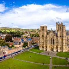 Aerial view of Wells Cathedral in England, with its Gothic architecture, green lawns in front, surrounding historic buildings, and a backdrop of trees and rolling hills under a partly cloudy sky.