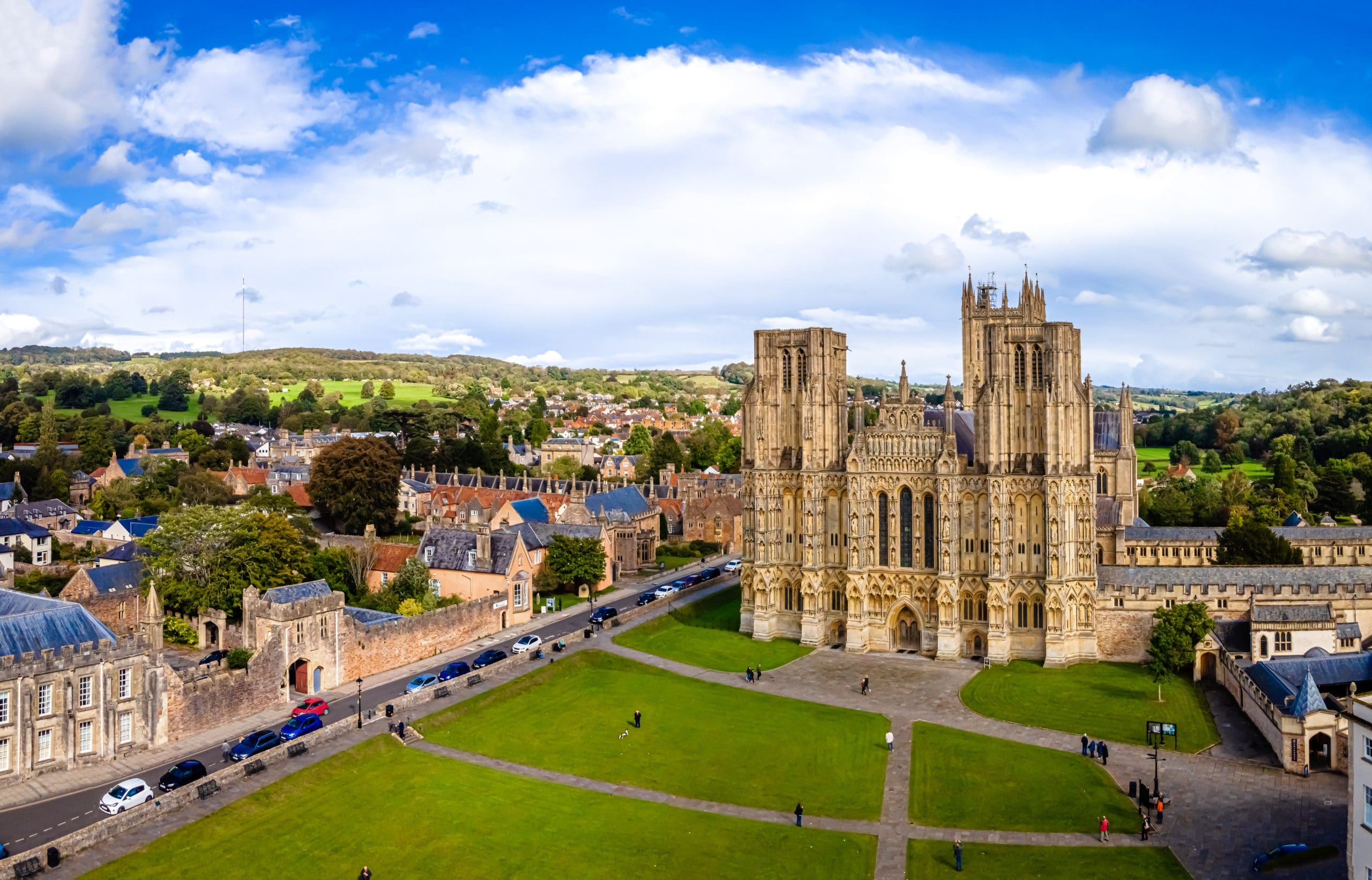 Aerial view of Wells Cathedral in England, with its Gothic architecture, green lawns in front, surrounding historic buildings, and a backdrop of trees and rolling hills under a partly cloudy sky.