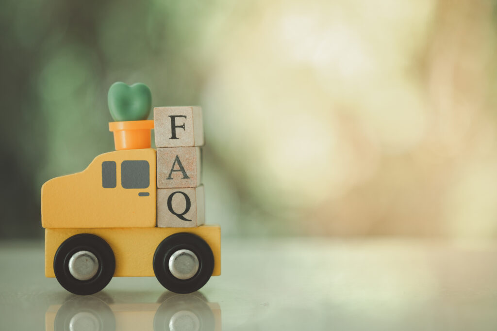 A small yellow toy truck carries three wooden blocks stacked vertically, each with a letter spelling "FAQ." A green heart shape sits on top of the blocks. The background is softly blurred.