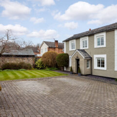A two-story gray house with white trim and a gabled porch, set beside a large paved driveway and a well-kept lawn, with trees and neighboring houses in the background under a partly cloudy sky.