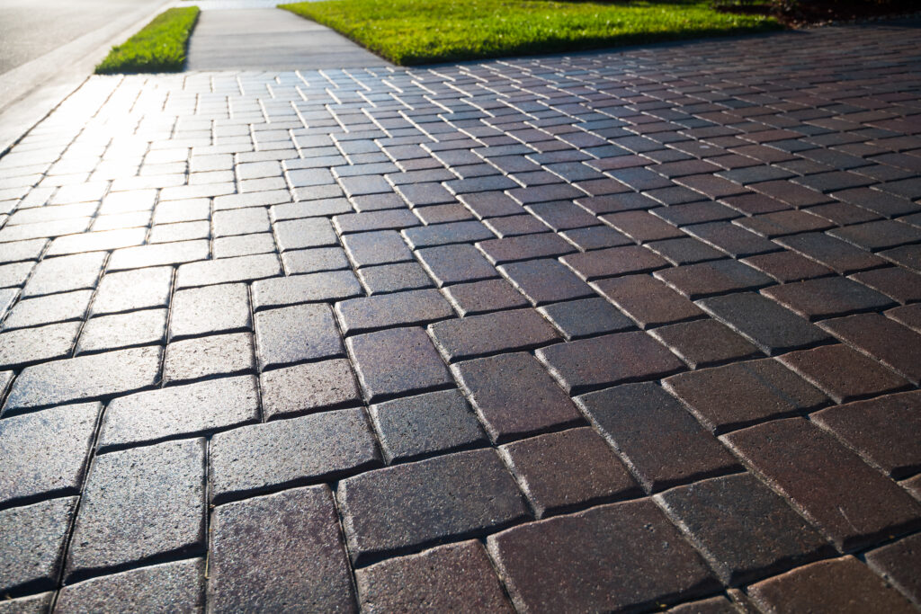 Close-up view of a clean brick or stone paved sidewalk with sunlight reflecting on the surface, next to a strip of green grass.