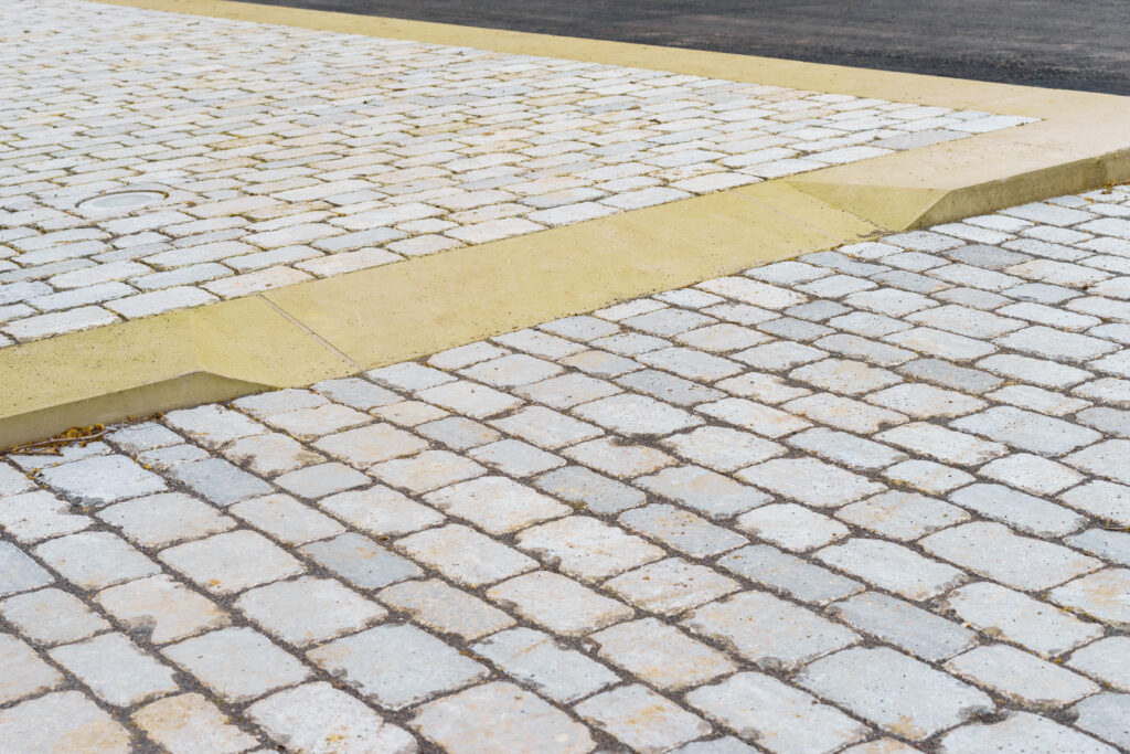 A smooth, wide curb ramp connects a cobblestone sidewalk to a paved street, providing accessible entry between the two surfaces. The stone blocks are light-colored and neatly arranged.