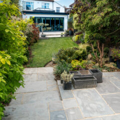 A landscaped backyard with a stone patio, potted plants, lush green lawn, trees, and a modern house with large glass doors in the background.
