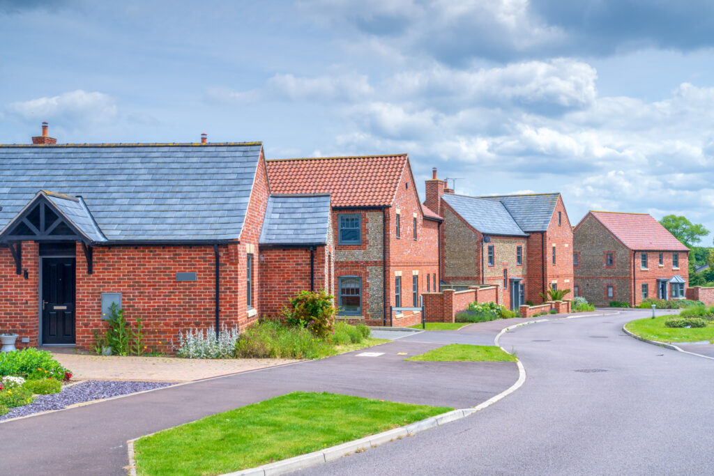 A row of modern brick houses with tiled roofs lines a quiet, curved street under a partly cloudy sky, surrounded by well-kept lawns and greenery.