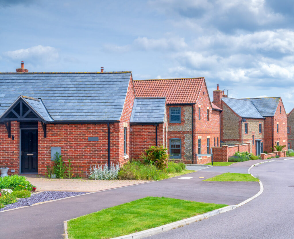 A row of modern brick houses with slate roofs lines a quiet, curved residential street under a partly cloudy sky. Manicured lawns and small gardens are visible in front of the homes.