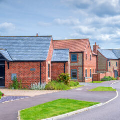 A row of modern brick houses with slate roofs lines a quiet, curved residential street under a partly cloudy sky. Manicured lawns and small gardens are visible in front of the homes.
